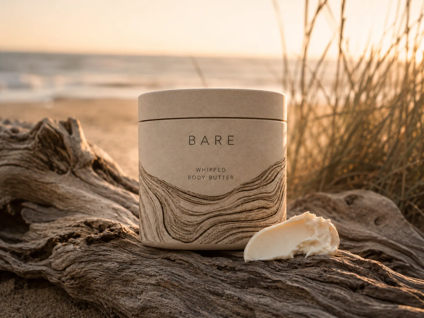 Bare whipped body butter jar with driftwood-grain packaging, styled on driftwood at golden hour by the sea